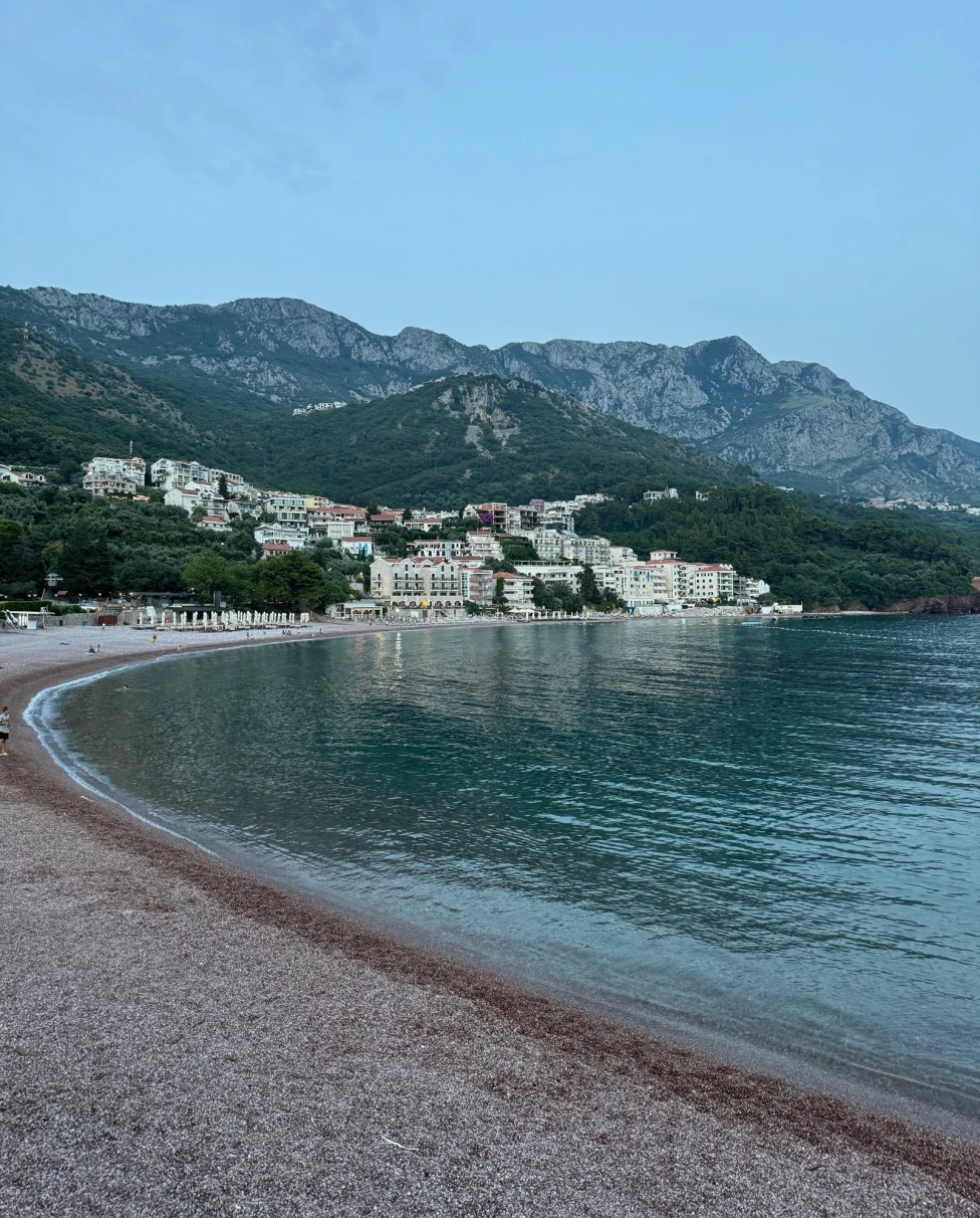 A scenic view of calm waves washing onto a beach with a coastal town in the disance and mountains at Kotor Bay, Montenegro.