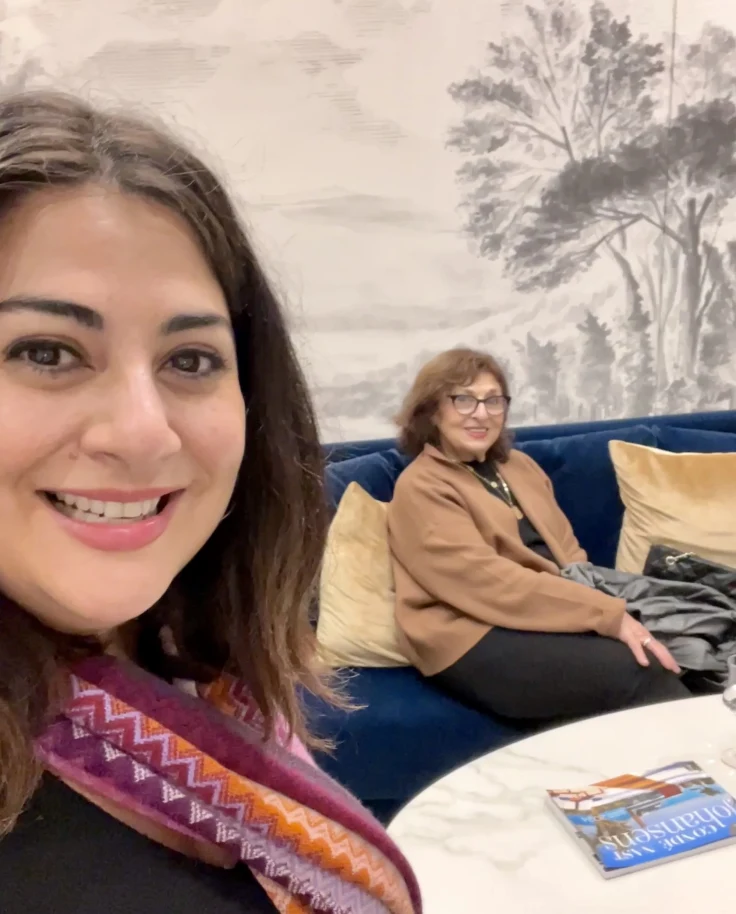 Jennifer posing for a selfie with a person in the background sitting on a blue couch near a white table.
