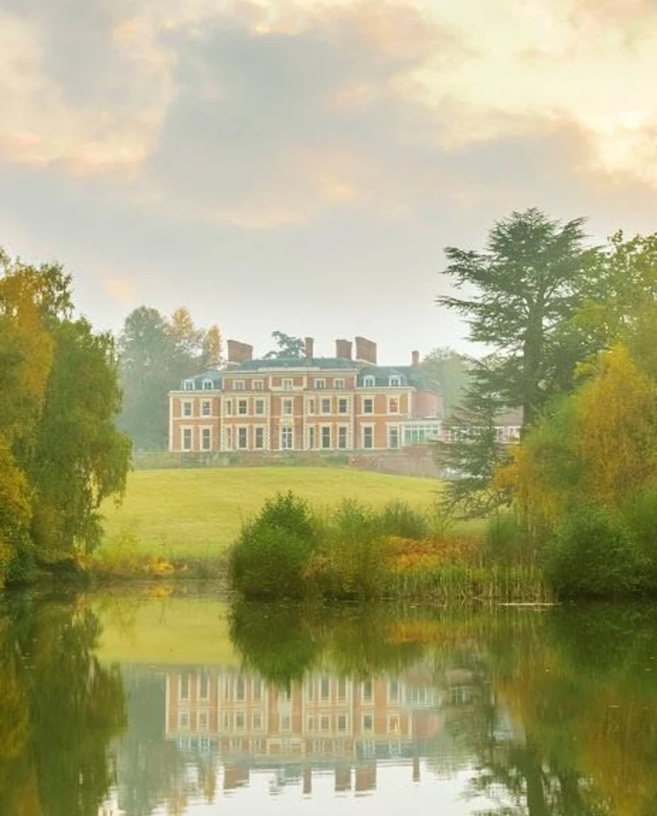 A view of Heckfield Place, a large red building with green roofs, in the distance surrounded by green grass, trees and a reflective body of water.