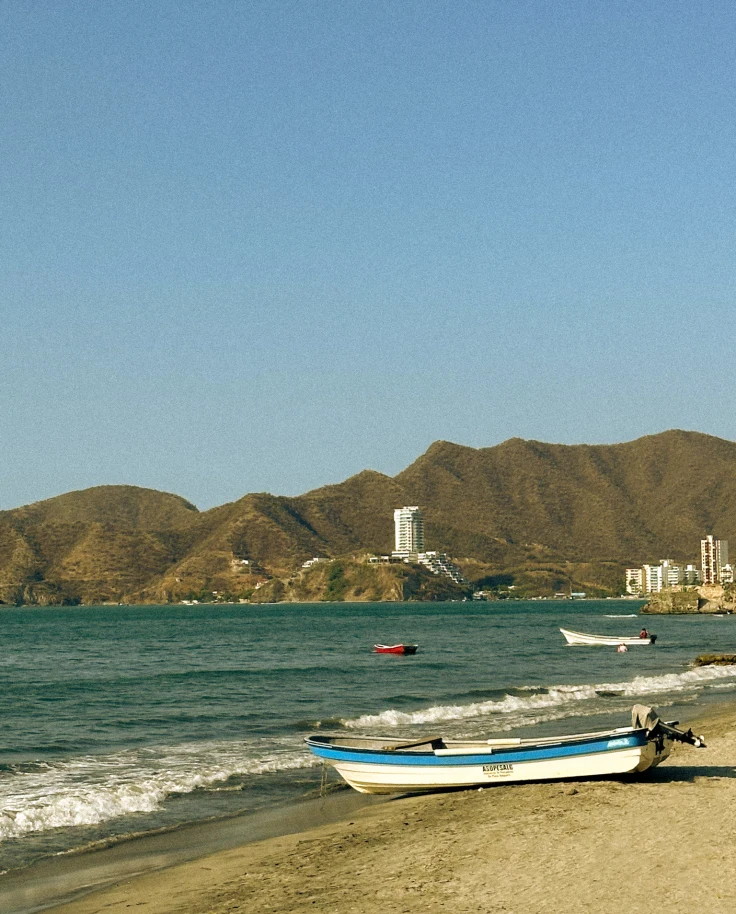 A view of a small boat on a sandy beach shore with the blue water, two boats, city shoreline and brown mountains in the background.
