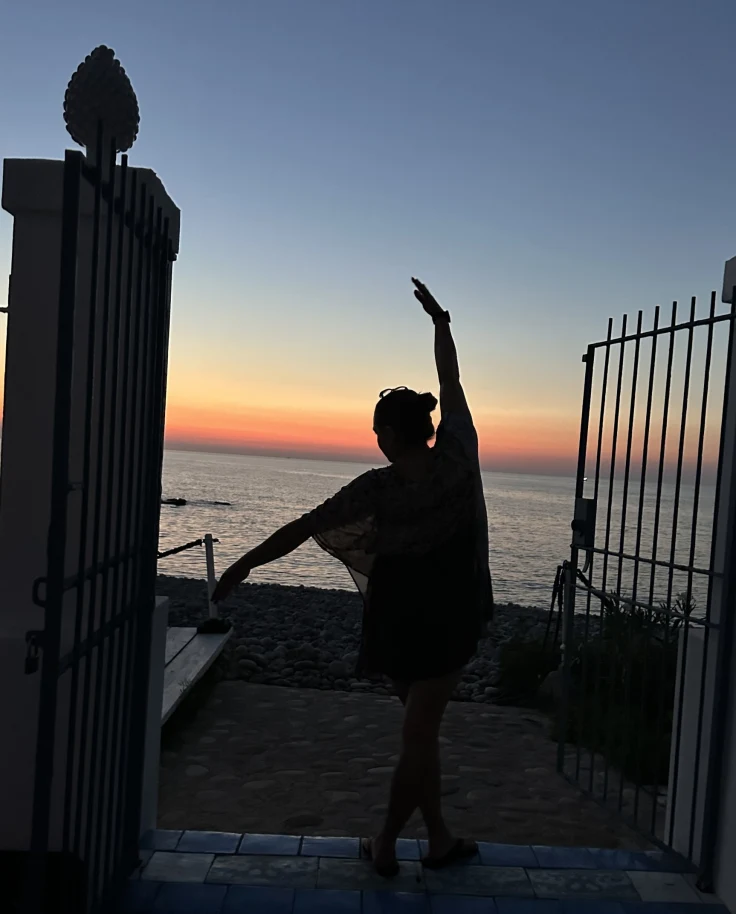 Image of April Bueno posing between two pillars and an open gate at sunset in front of the ocean