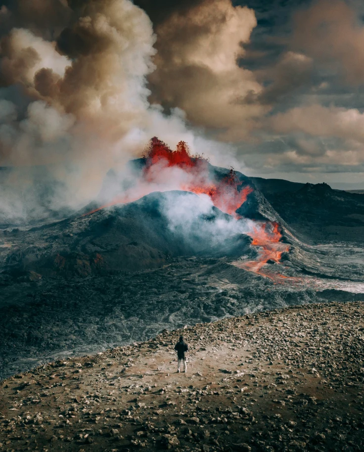 A person standing on rocky terrain with an active volcano and eruptions of smoke in the background.