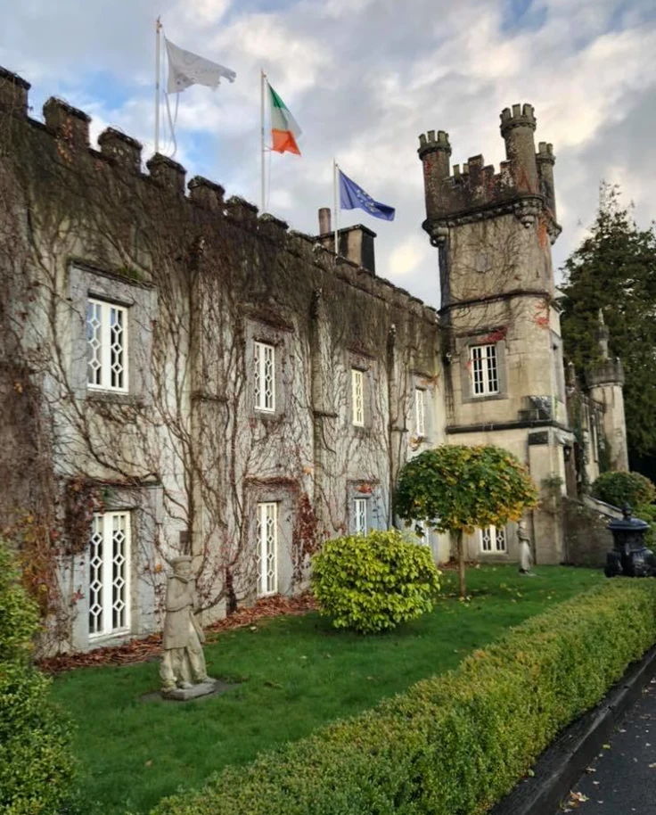 A view of a stone castle's exterior with three flags perched on top, with a green lawn and trees surrounding it on a sunny day.