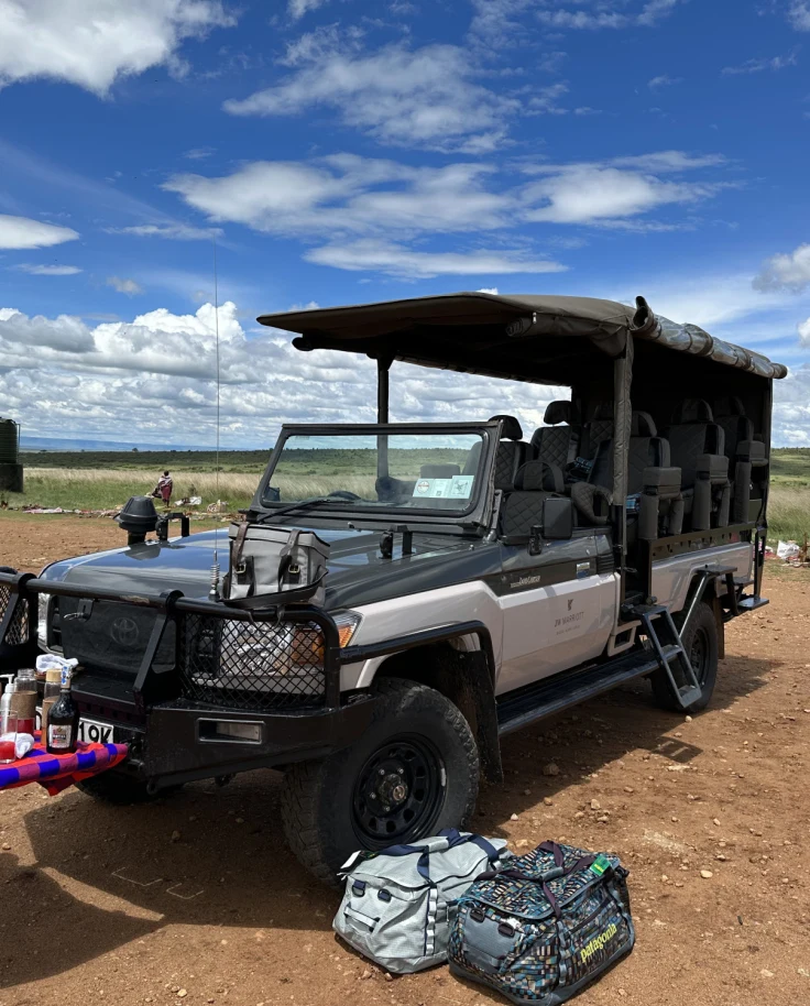 A covered Jeep with open sides parked in the middle of a dirt road, with a grassy field behind it, with a makeshift table extended in front of it covered in plates of food and condiments.