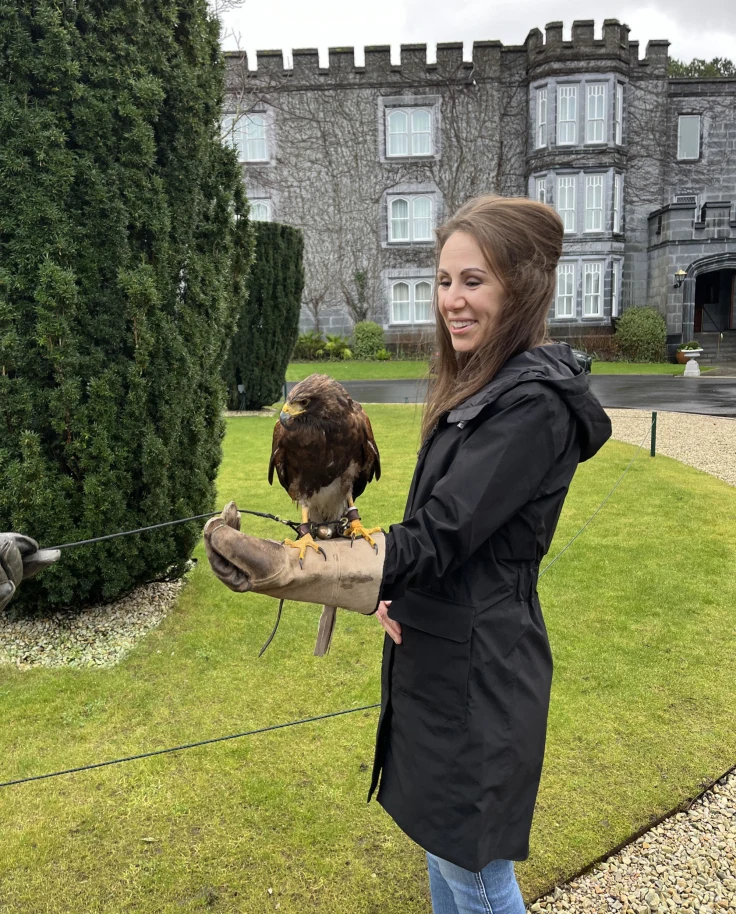 Megan standing in a jacket and blue jeans with a bird on their arm and castle in the background surrounded by green trees and grass.