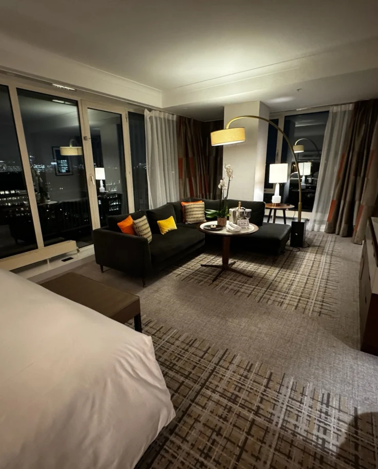 A hotel room with a white bed and couch area, with floor-to-ceiling windows overlooking the city at night.