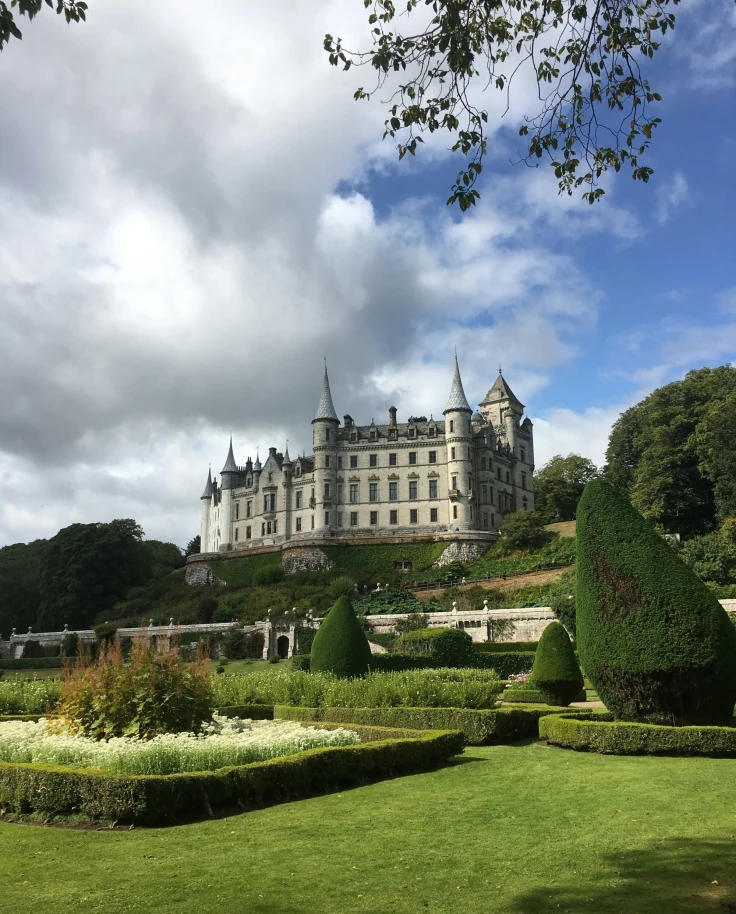 A castle sits atop a hill surrounded by manicured gardens on a cloudy day.