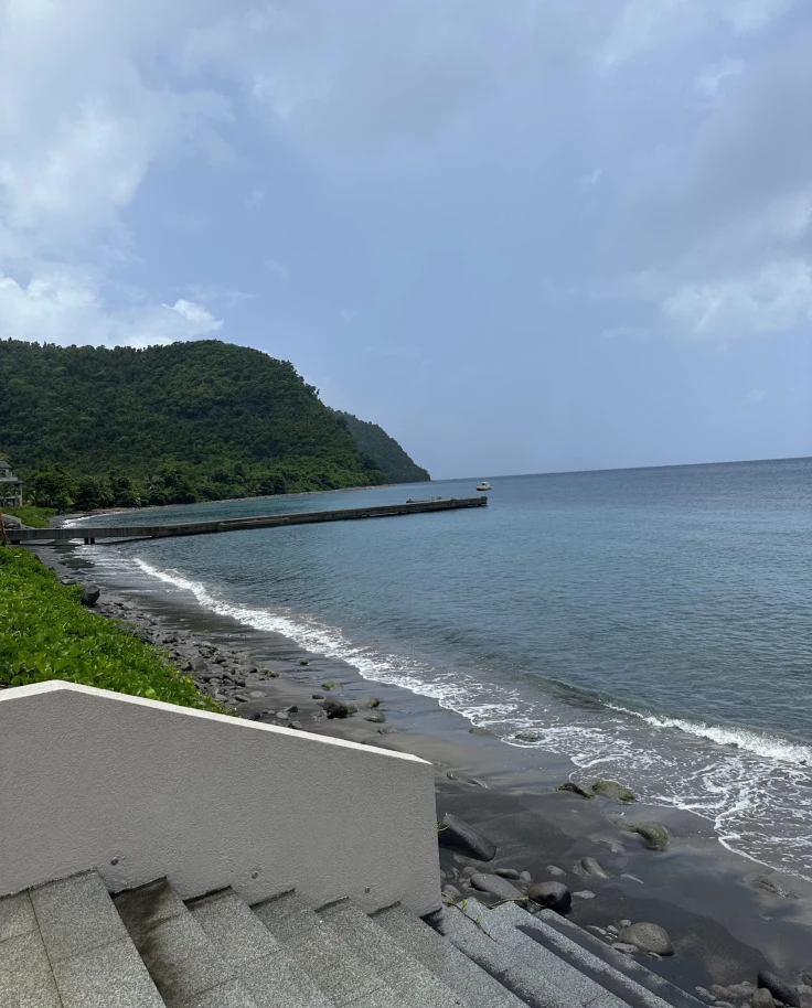 A view of white stairs leading to a dark sandy beach and blue sea. There is a green mountain in the background.
