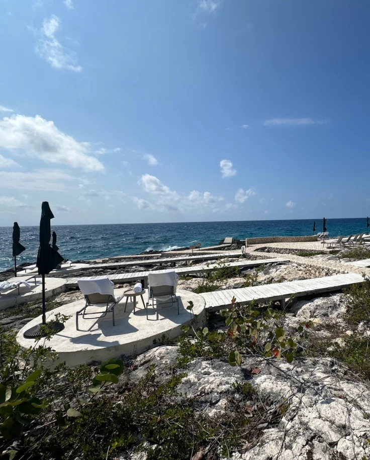 A beautiful view of a sandy beach with white boardwalks leading to a seating area complete with two lawn chairs and an umbrella overlooking the blue water.
