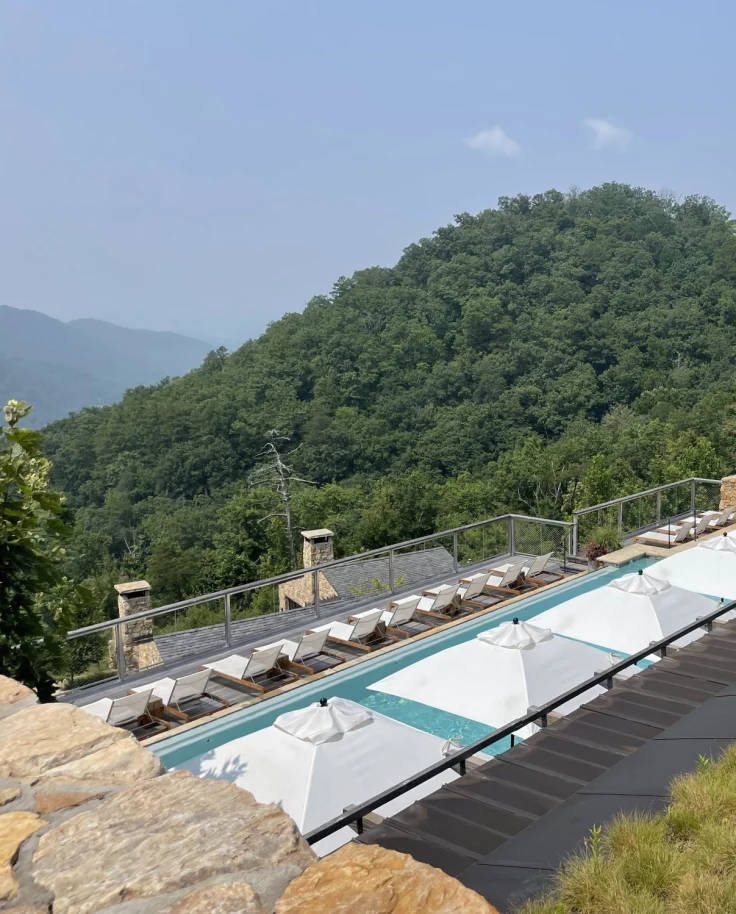 A top down view of a swimming pool deck complete with rows of white umbrellas and lounge chairs overlooking a lush, green mountain.