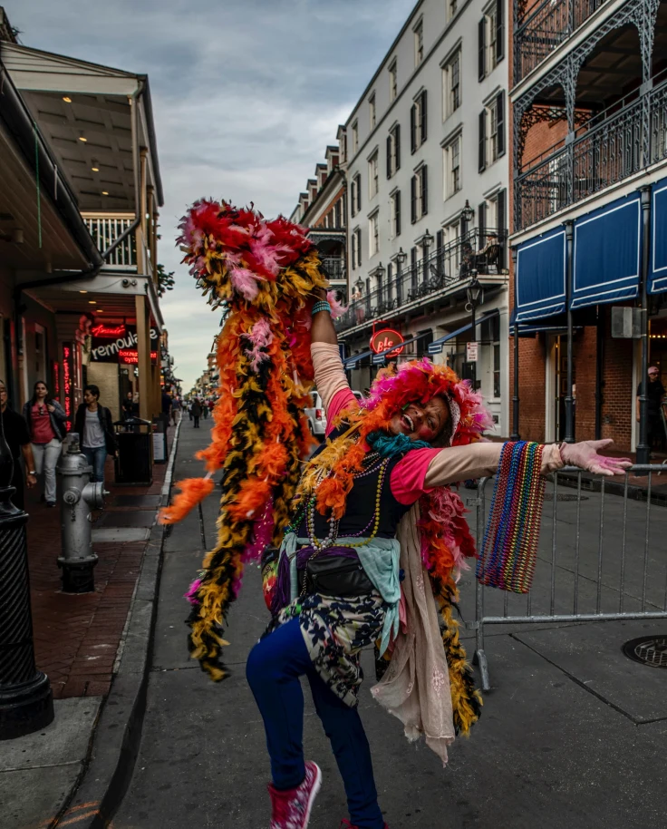 A woman adorned in flowers with her hands spread open posing in front of buildings outside in New Orleans.