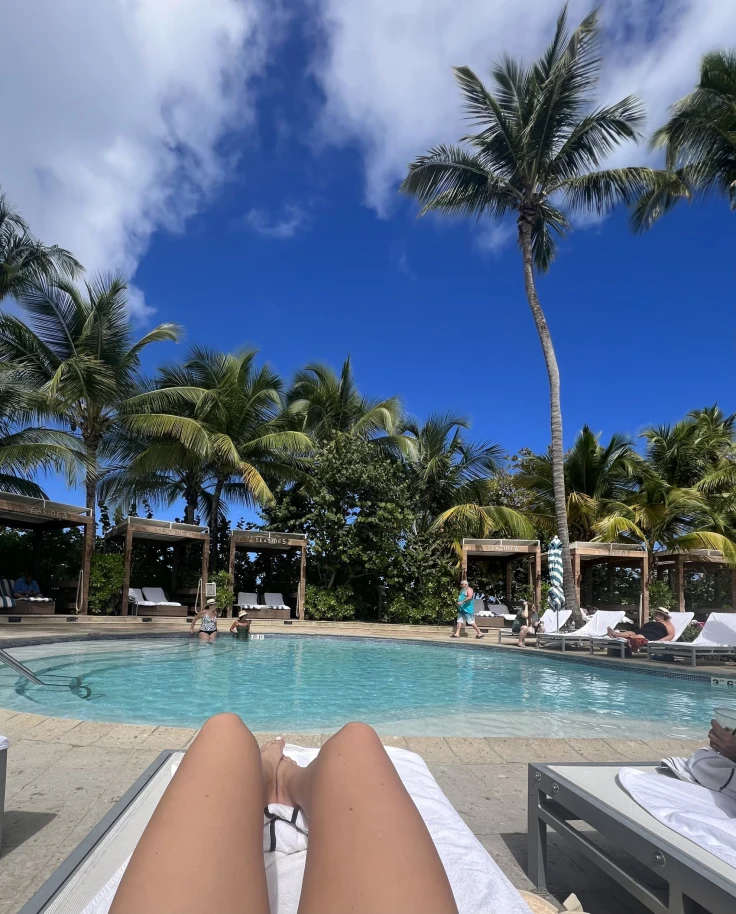 A person tanning their legs on a lawn chair by a swimming pool surrounded by palm trees.