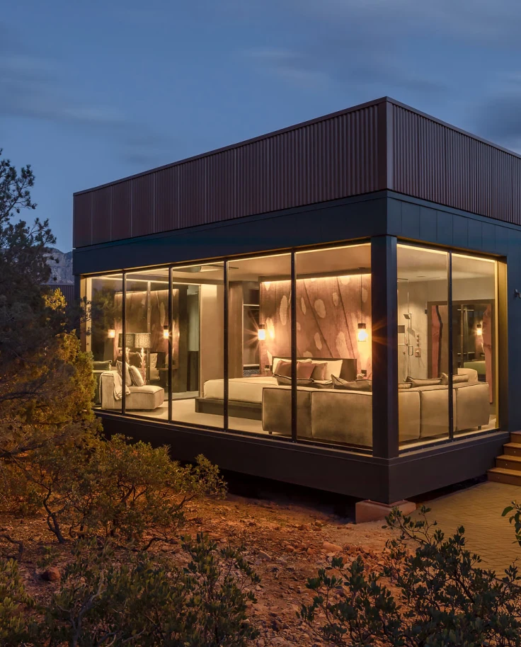 The exterior of a room at Ambiente in the middle of nature at dusk, with floor-to-ceiling windows showcasing a bed and couch inside.