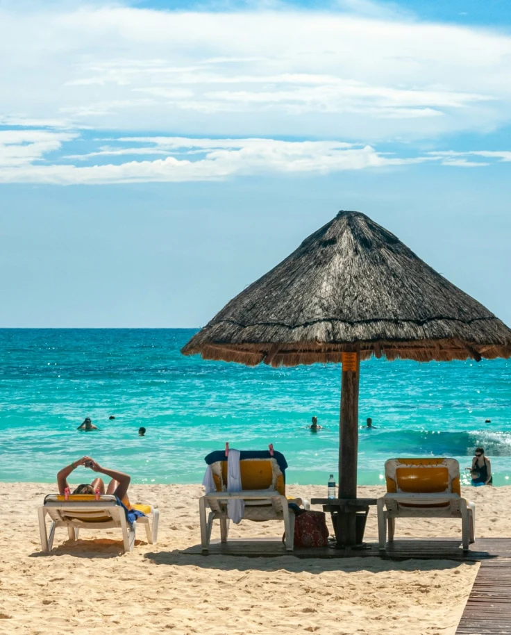 A beach with beach chairs and a thatched umbrella looking out at a turquoise ocean on a sunny day near the Cancun Mexico Grand Mayan Riviera Maya.