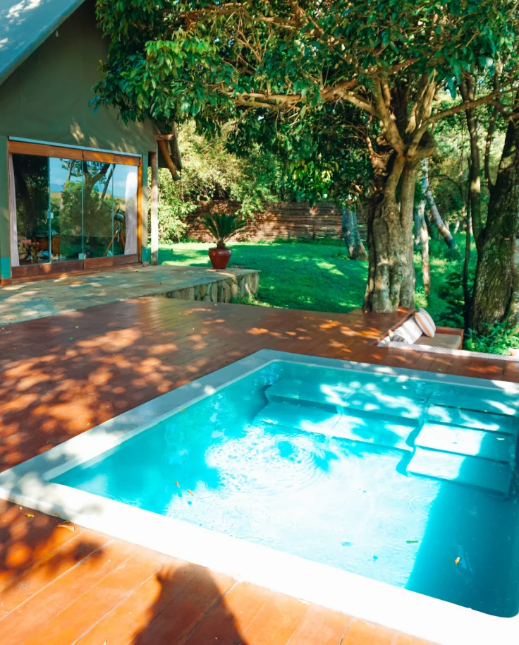 A patio with a pool, trees, wood fooring and a house with potted plants around it on the left side of the image.