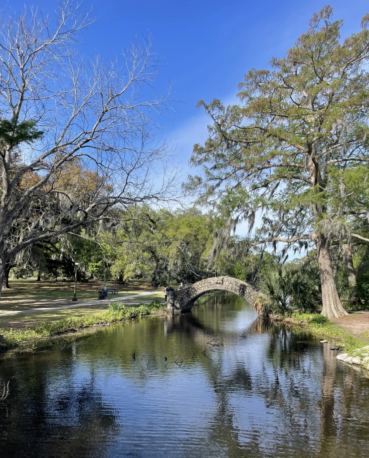 An image of a creek with a stone arch bridge in the distance, surrounded by tall trees with green leaves and green grass beneath a blue sky.