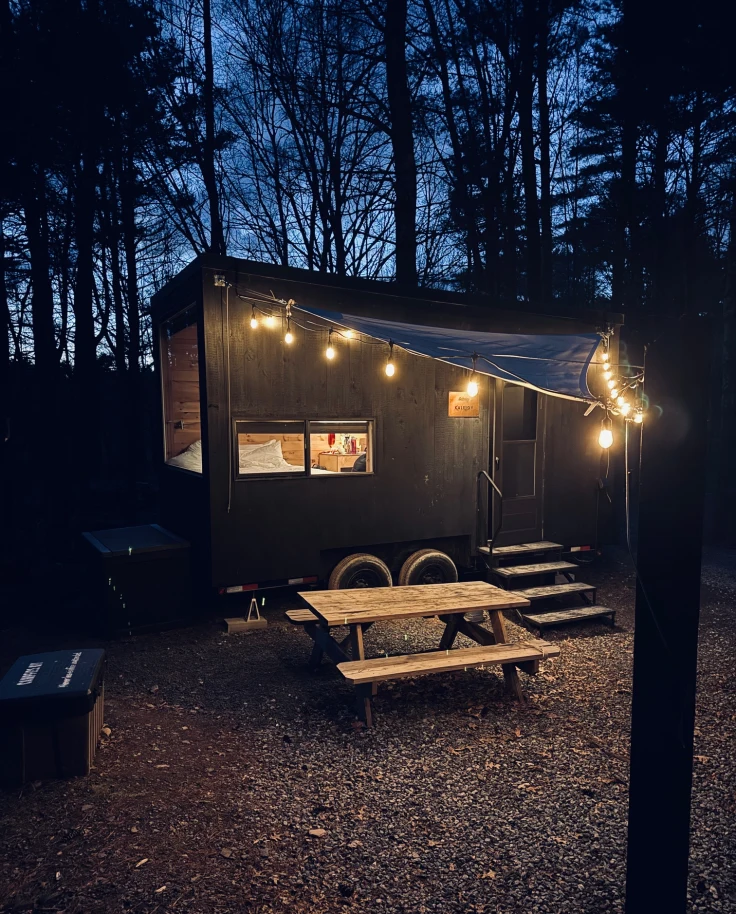 A trailor in the woods with a string of lights illuminating it, and a picnic table in front at night.
