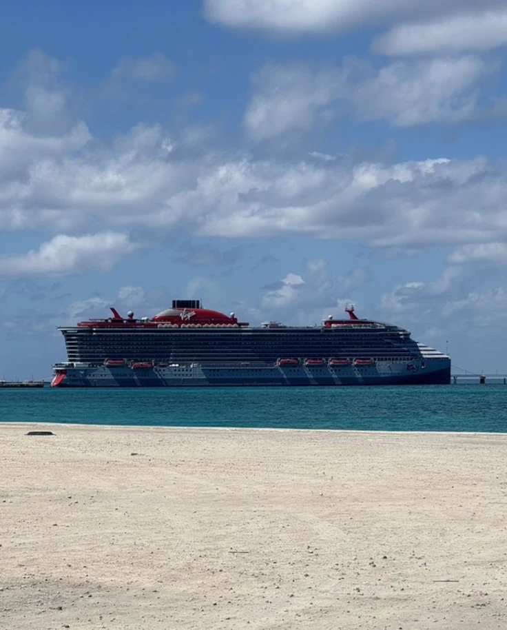 A view from a beach of a large Virgin Voyages cruise ship sailing