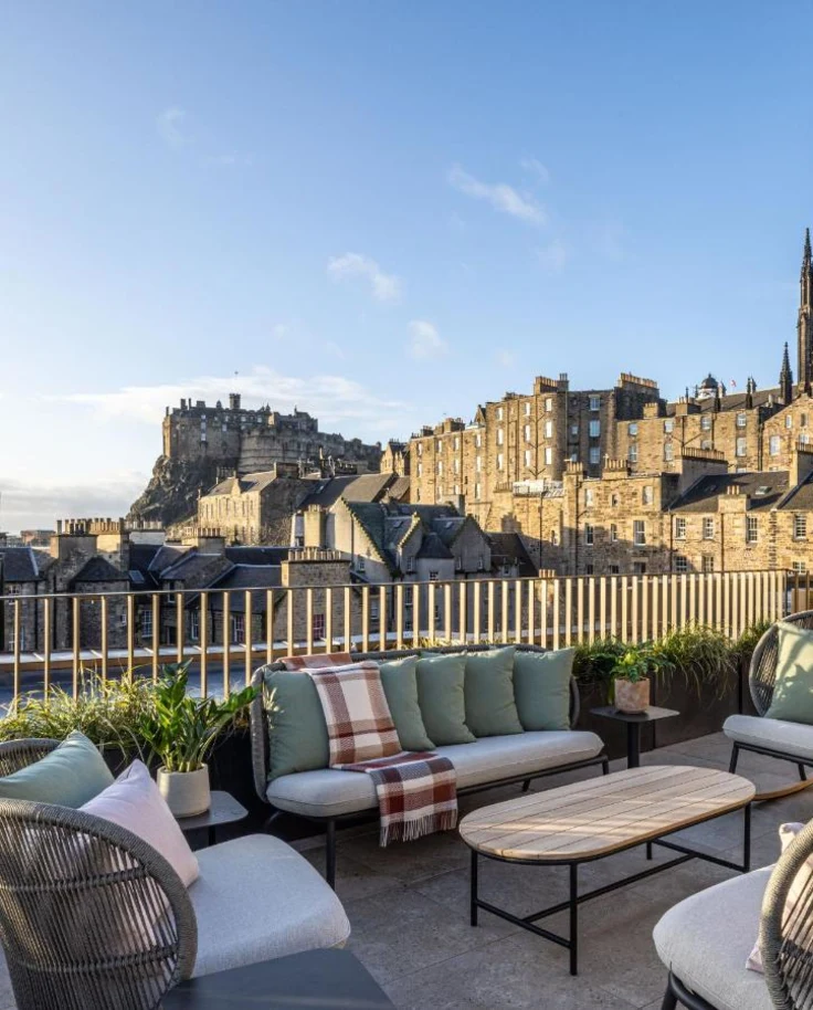 A view of an outdoor patio with couches, a table and a stone castle in the background surrounded by stone buildings and a church tower.