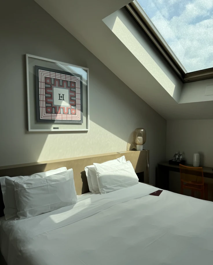 A photo of a white linen bed underneath an A-frame roof with a window letting light in from it onto the bed.