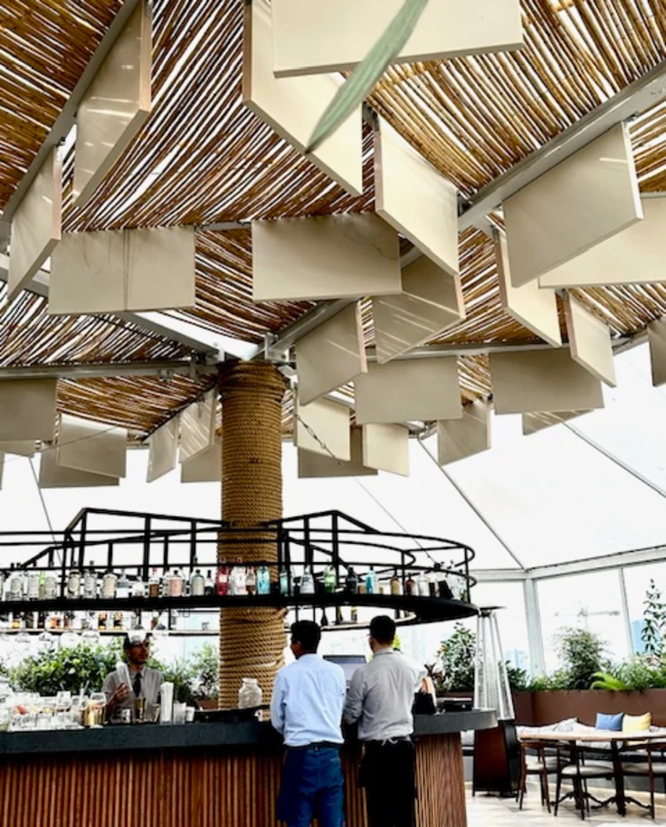 This image depicts a view of two people standing with their backs turned toward a bar beneath intricate ceiling designs and wood panels. There is liquor displayed in front of them and seating in the surrounding areas around them.