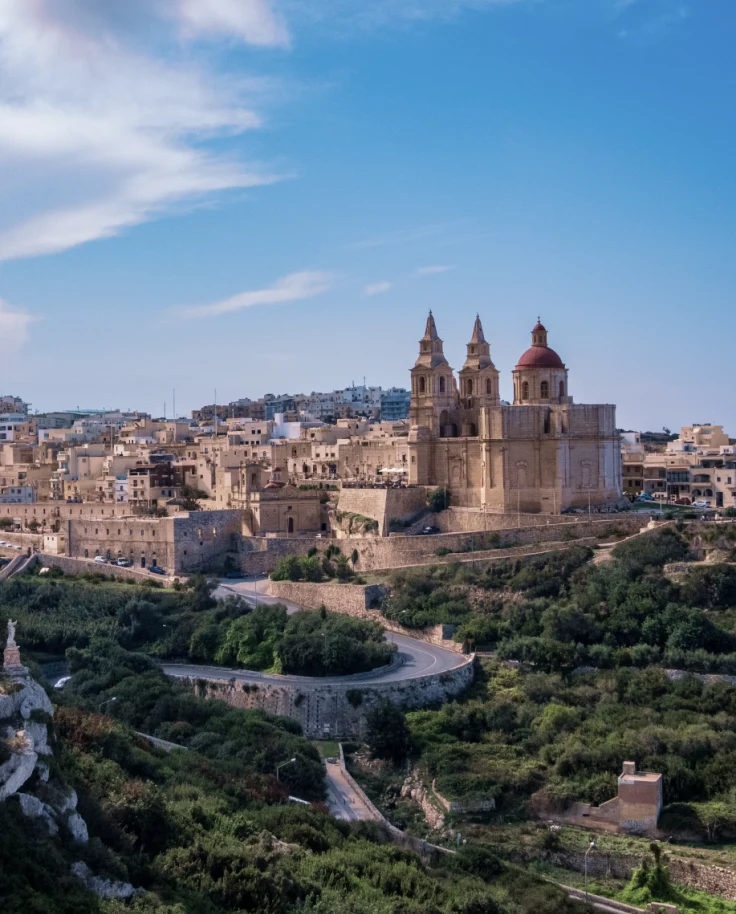 A beautiful aerial view of stone buildings tucked into a coastal array of rocks, trees and winding roads in Malta.