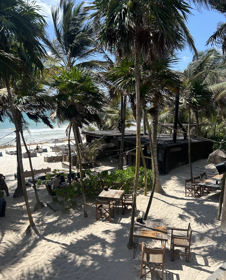 A view of the beach at Be Tulum hotel with wooden chairs and tables and palm trees