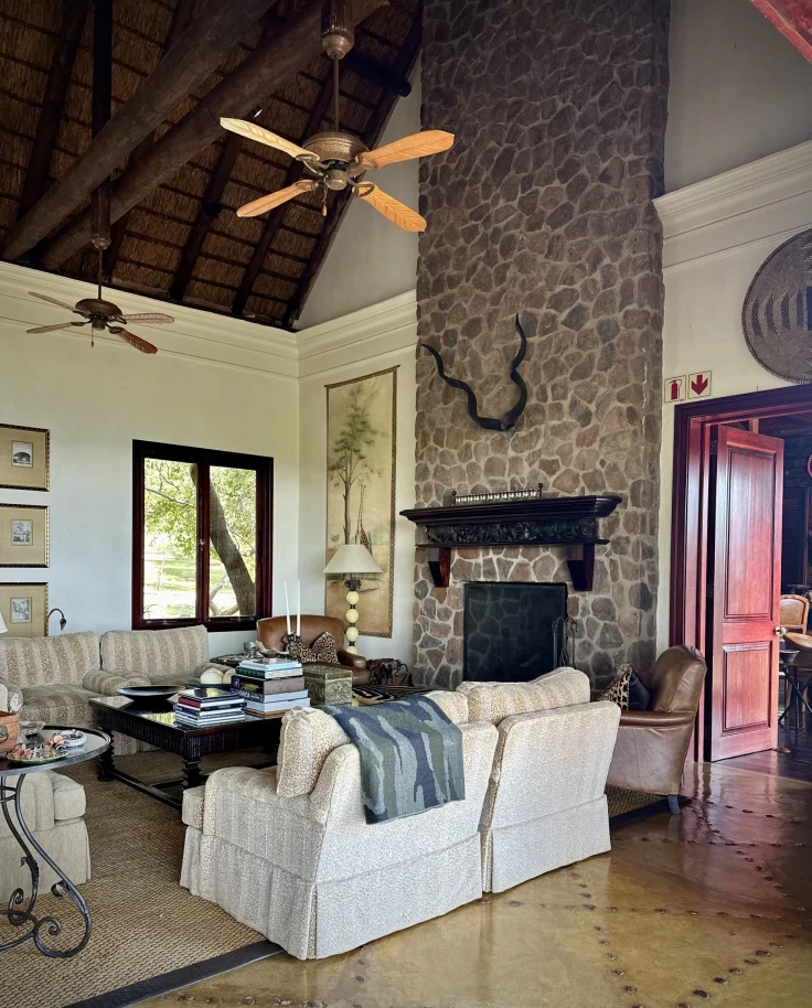 An interior view of a room with a large stone fireplace, mounted antlers, a ceiling fan, striped couches, tables, lamps, art, tiled flooring and red doors beneath a wood beamed ceiling.