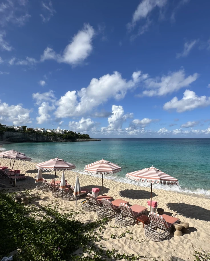 Loungers and umbrellas line the shore as gentle waves lap the shore on a sunny day dotted with clouds.