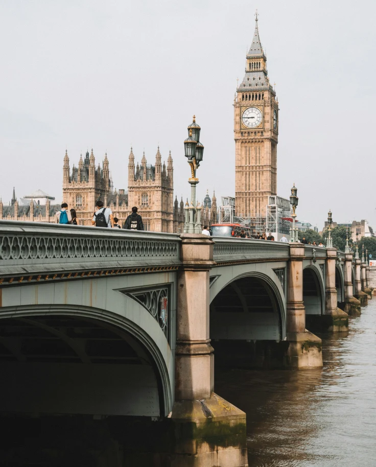 Tourists walking along a London bridge over rushing water under a gray sky.