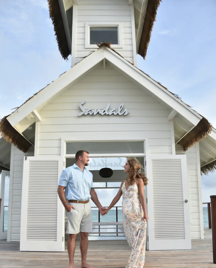 Couple in beach attire in front of a white Sandals building with a view of the ocean in the background