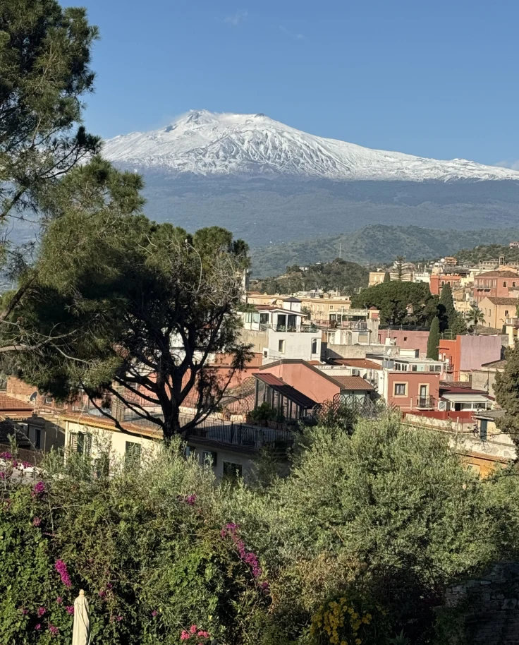 View of a town in Sicily with green trees and picturesque buildings and a snow-capped mountain in the background