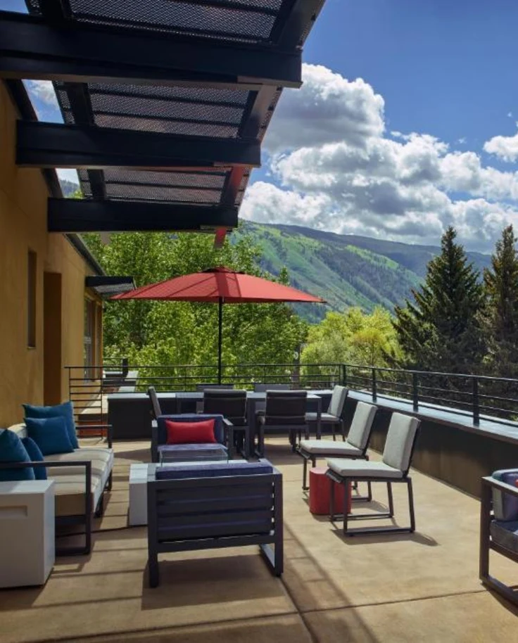 This image depicts a hotel terrace at The Limelight Aspen with various options for lounge seating and umbrellas. There is a stunning view of tree covered mountains and a cloudy blue sky in the background.