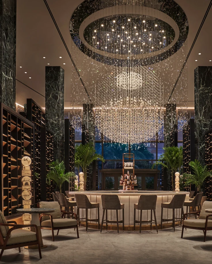 A view of a luxury hotel interior that is dimly lit with large pillars on either side, slatted wood walls, a circular table and chairs surrounding it with a large oval chandelier hanging from the ceiling.