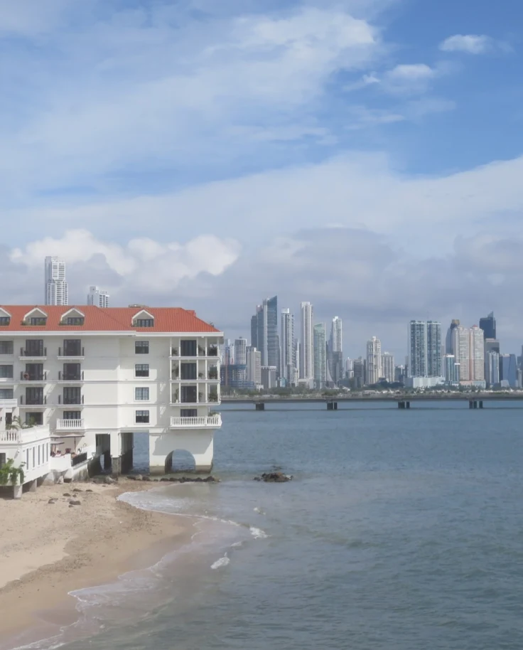 The Sofitel Legend Casco Viejo Panama City from the exterior - a white building on the water with a red roof and the city skyline in the background - Suzanne Tomlin