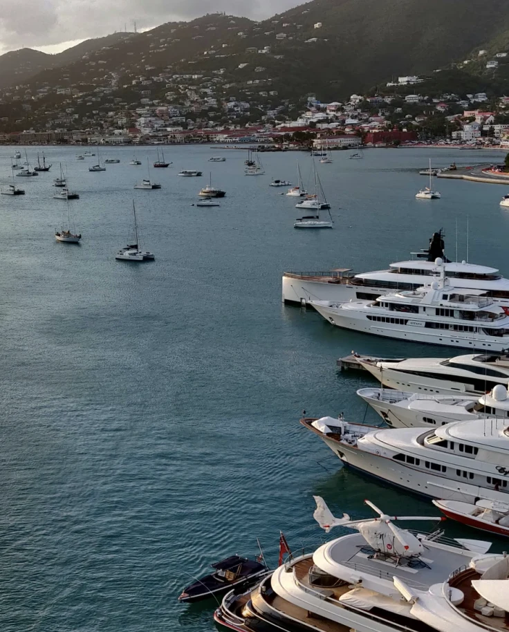 A view of yachts anchored in a harbor with sailboats and buildings tucked into a mountain in the background.
