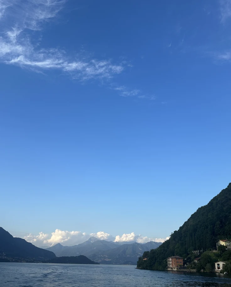 A view from the ferry on the lale with mountains, low puffy white clouds and a big blue sky during the day.