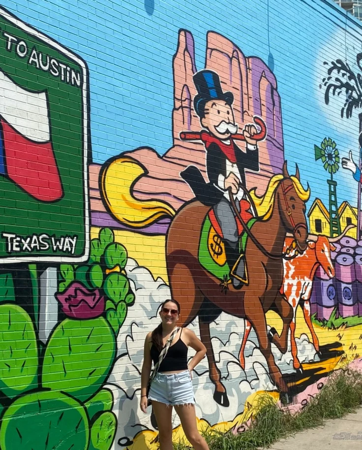 A painted mural with various images including a Texas flag, cactus, horse, people, rocks and more with a woman posing in front of it.