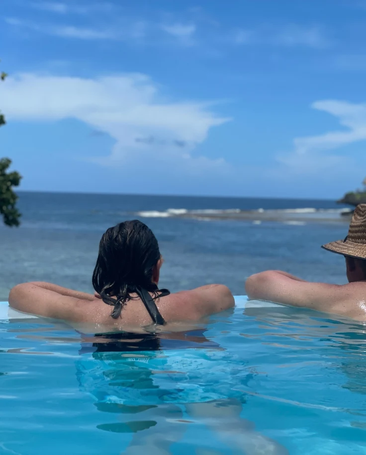 A couple in a swimming pool resting on the edge facing the ocean