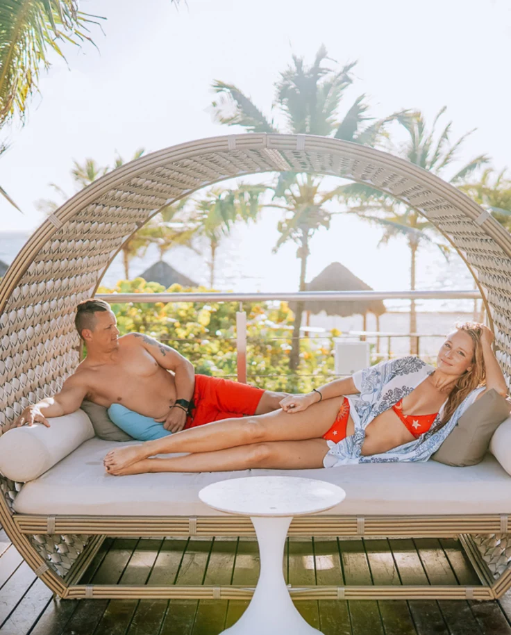 A couple lounging in a cabana on a wooden patio with white umbrellas and palm trees in the surrounding areas.