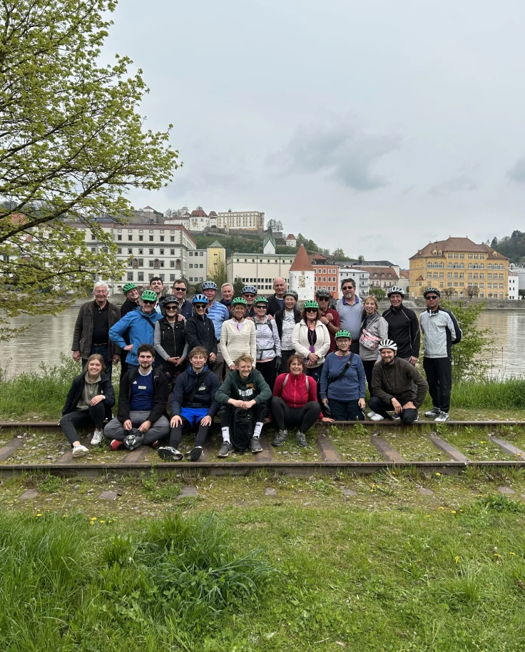 A group photo of travelers, about half of whom are wearing bicycle helmets, on the grassy by the Danube River on a cloudy day.