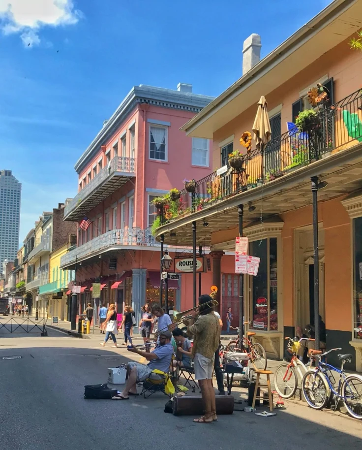 A group of people playing musical instruments on the road next to yellow and pink buildings with flags, metal balconies and plants.