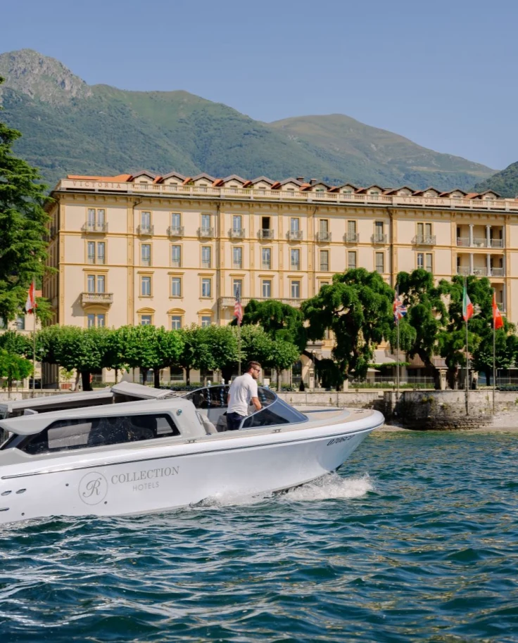 A white boat on Lake Como in front of a luxurious yellow hotel with green trees and mountains.