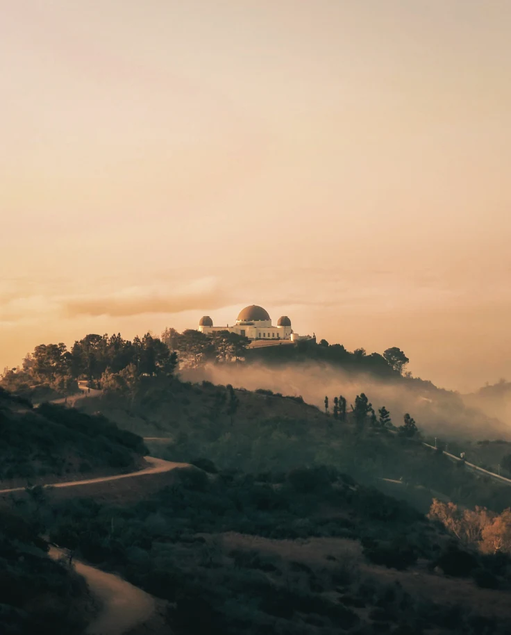 A view from one of the hotels near Griffith Observatory, featuring a large white building with a domed roof, on top of a hill covered in trees at sunset.