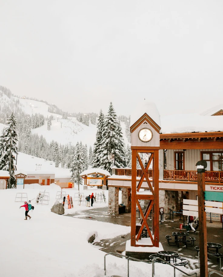 A view of a ski lodge, clock tower and people walking on the snowy terrain in the distance towards snowy pine trees and a ski hill that escapes into the fog.