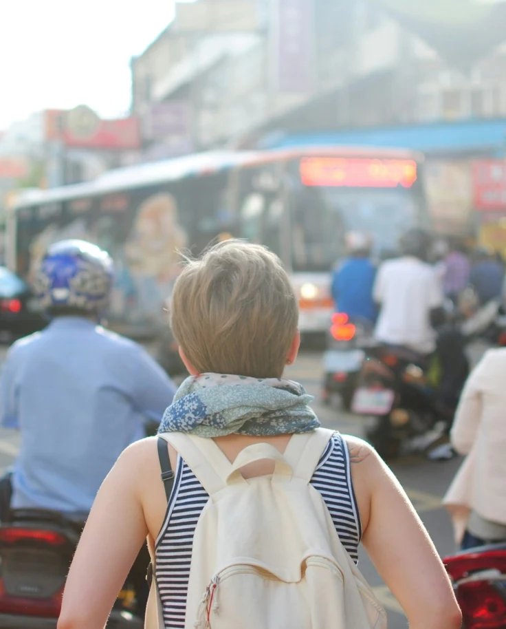 A girl wearing a striped top, scarf and white backpack walking solo down a busy street.