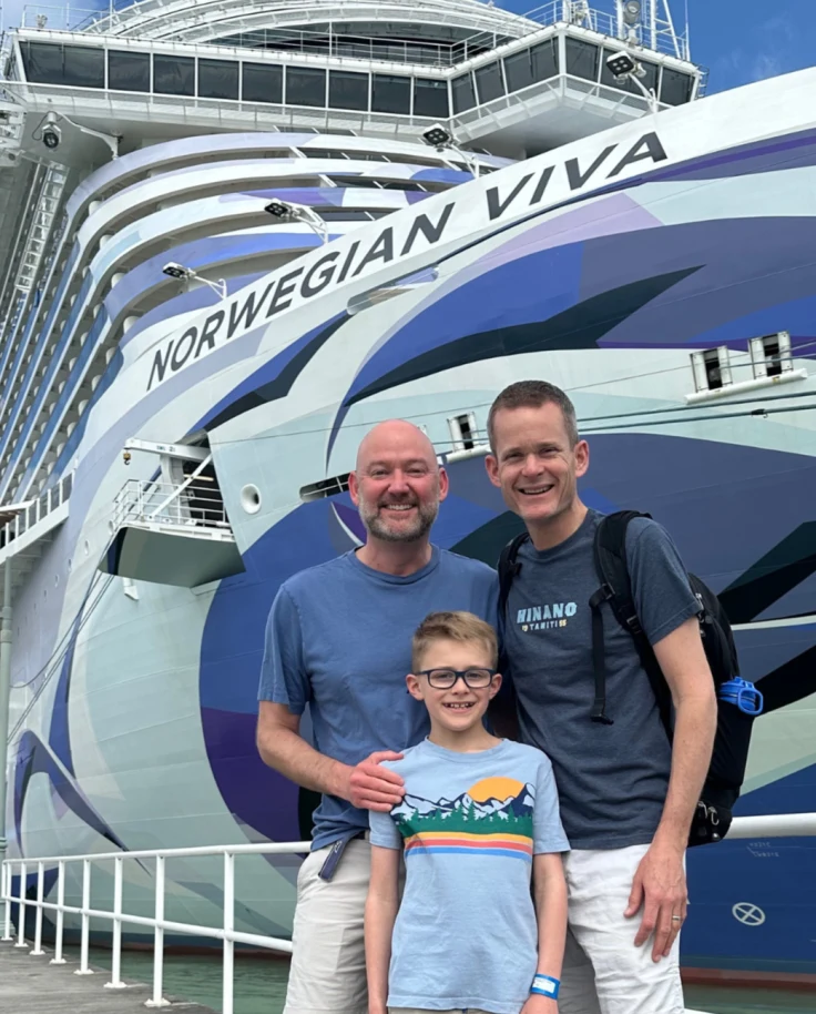 A father and two sons stand in front of a cruise ship.