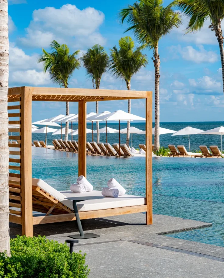 A swimming pool with wooden cabanas, white umbrellas and palm trees.