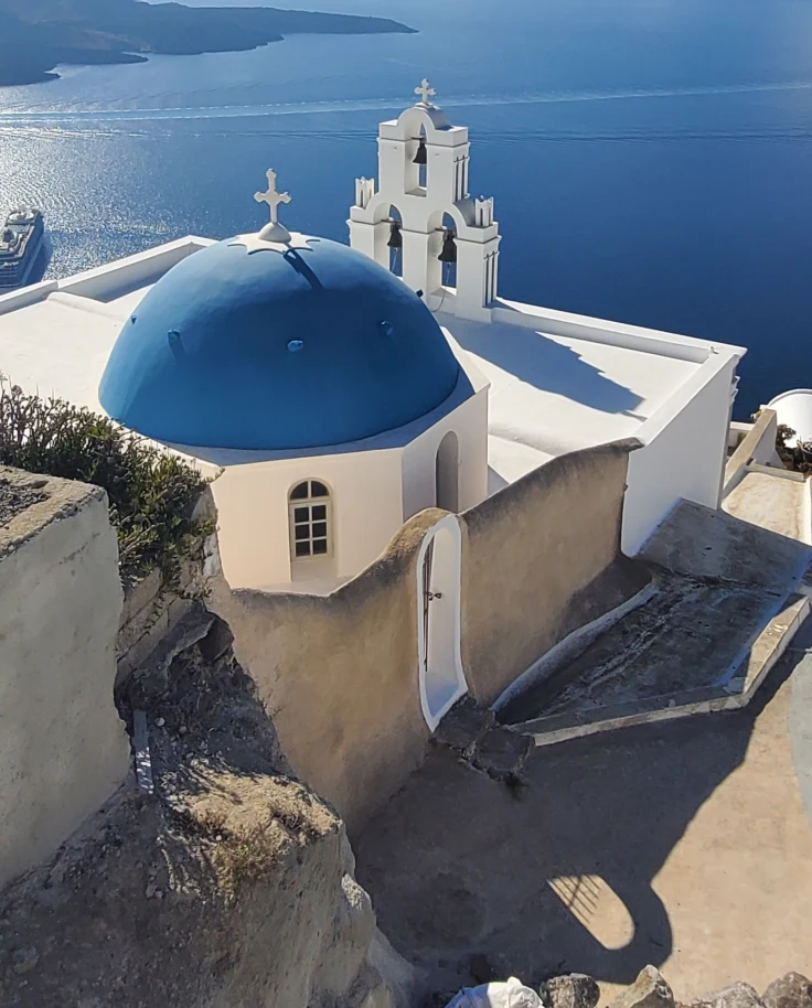 A view of the coastline of Santorini, Greece, with bright white buildings and the famous blue dome overlooking the ocean on a sunny day.