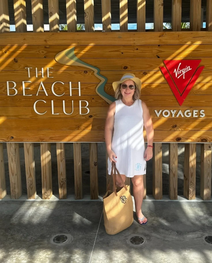 Shelly Maffia in a white outfit and sun hat standing in front of a wooden sign that says "The Beach Club - Virgin Voyages"